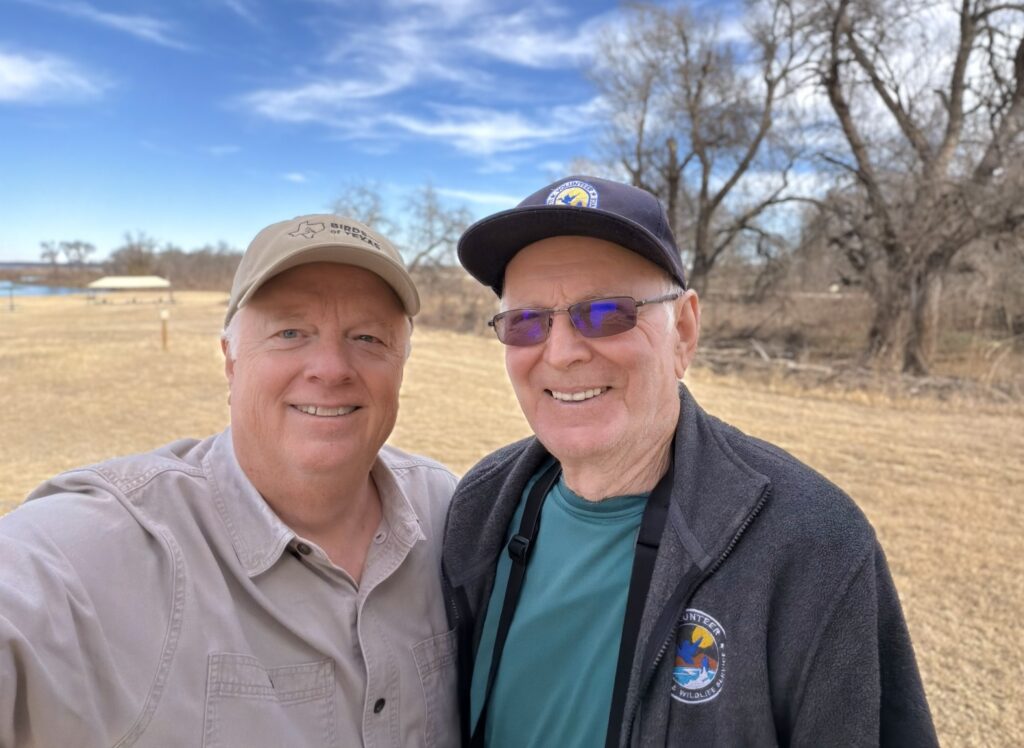 Two men smiling outdoors on a sunny day with trees in the background.